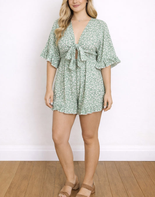 Woman wearing a green floral Tie Front Romper against a white wall.