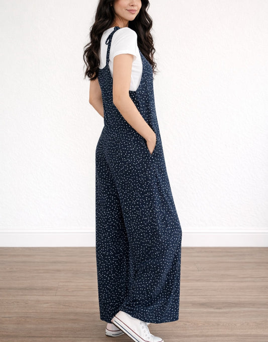 Side view of woman wearing navy Tie Shoulder Jumpsuit with white polka dots and white tee underneath on plain white background.