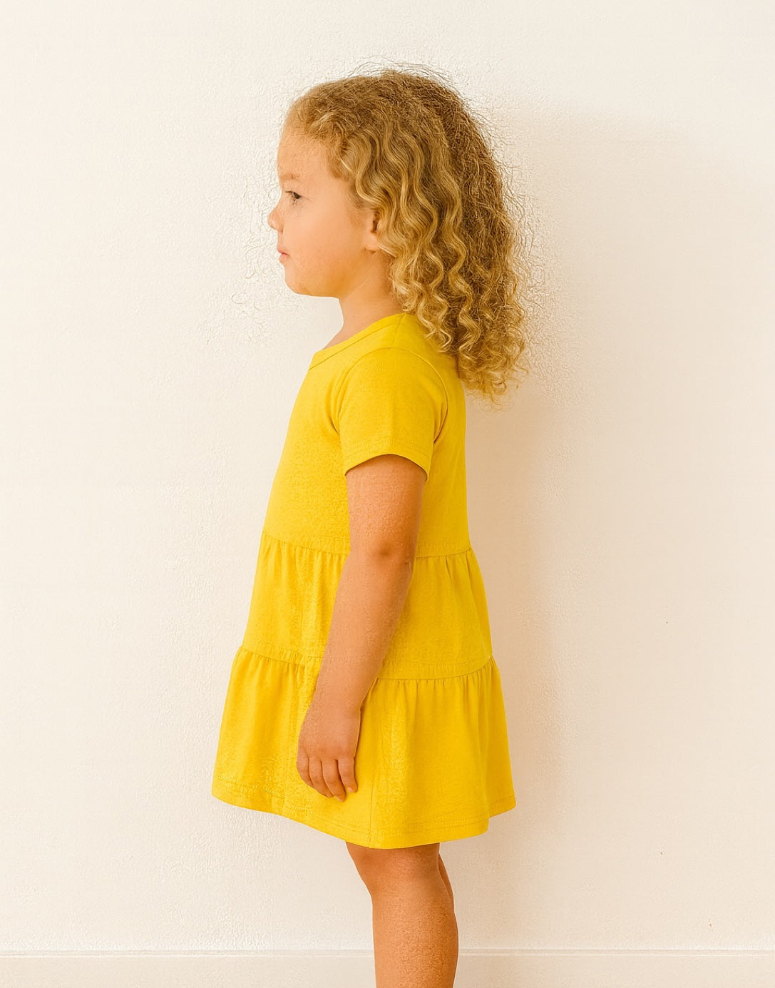 Side view of young girl wearing a yellow Ruffle Hem T-Shirt Dress against a white background.