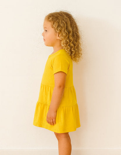 Side view of young girl wearing a yellow Ruffle Hem T-Shirt Dress against a white background.