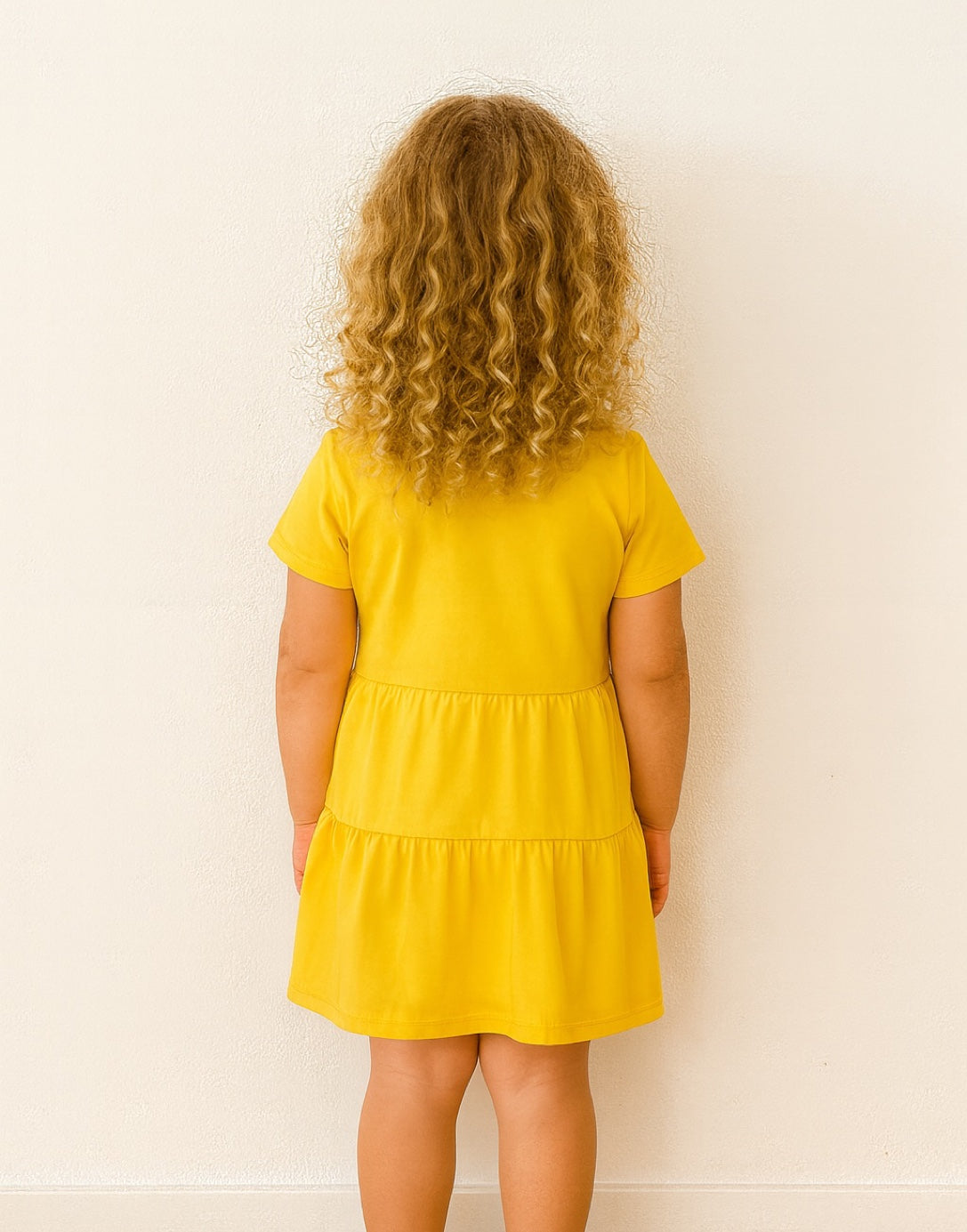Back view of young girl wearing a yellow Ruffle Hem T-Shirt Dress against a white background.