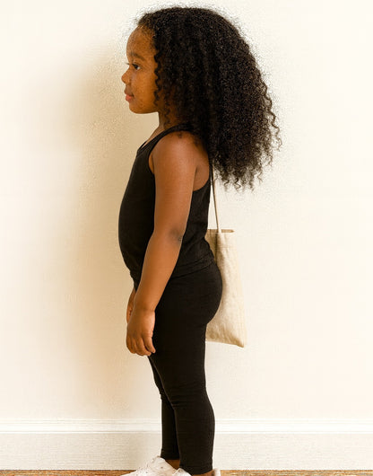 Side view of young girl in black tank top and leggings set holding a white bag against a plain background.