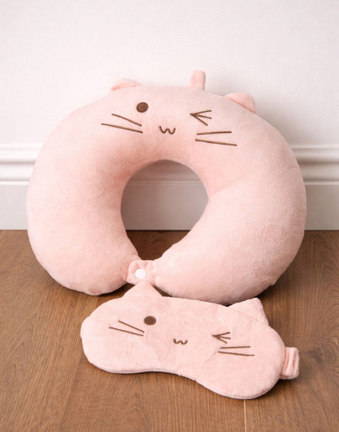 Pink cat-shaped neck pillow and eye mask on a wooden surface with a white wall background.