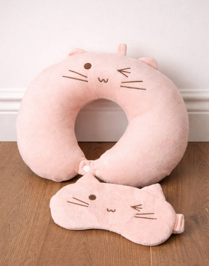 Pink cat-shaped neck pillow and eye mask on a wooden surface with a white wall background.