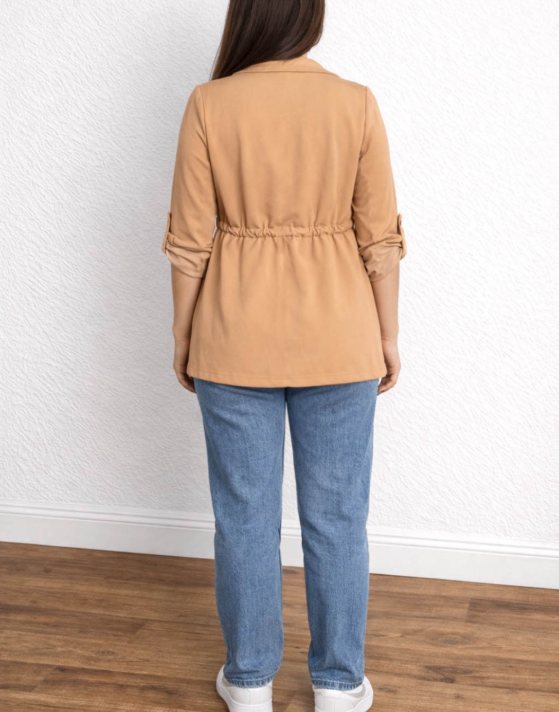 Back view of woman wearing a tan Waterfall Jacket, blue jeans, and white sneakers against a white wall.