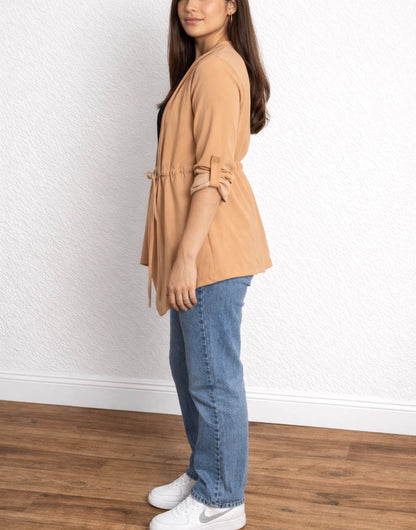 Side view of woman wearing a tan Waterfall Jacket, black top, blue jeans, and white sneakers against a white wall.